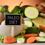 closeup of a signboard with the text paleo diet on a table full of different raw vegetables, a bowl with some chicken eggs and a chicken