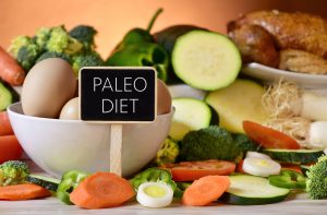 closeup of a signboard with the text paleo diet on a table full of different raw vegetables, a bowl with some chicken eggs and a chicken